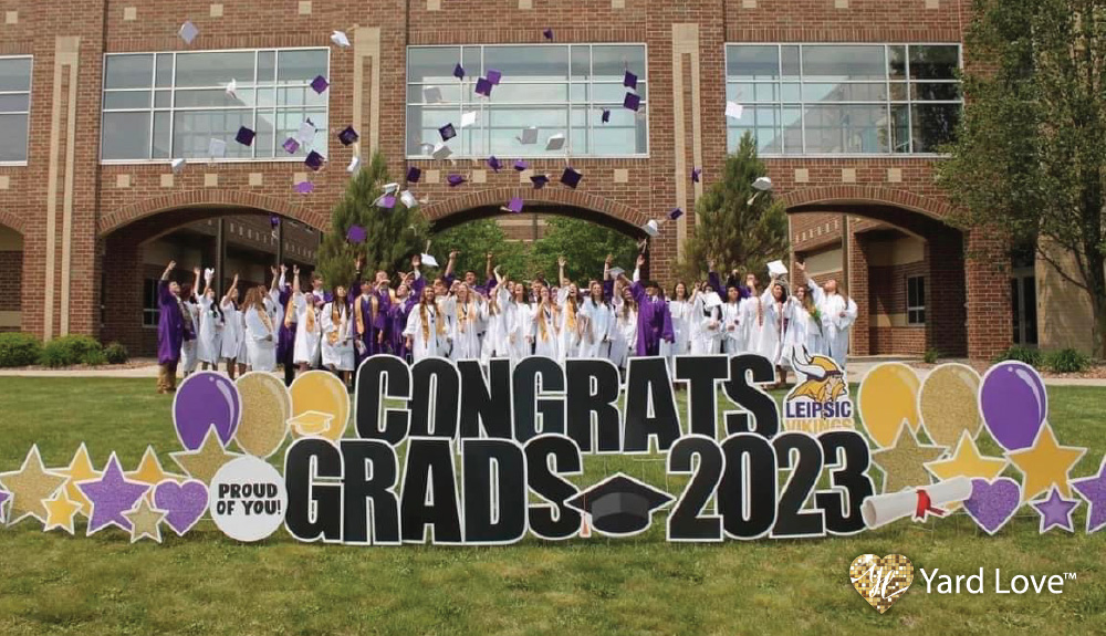 high school graduates in white and purple robes throwing caps into the air behind a Congrats Grads 2023 Leipsic Vikings Yard Love sign