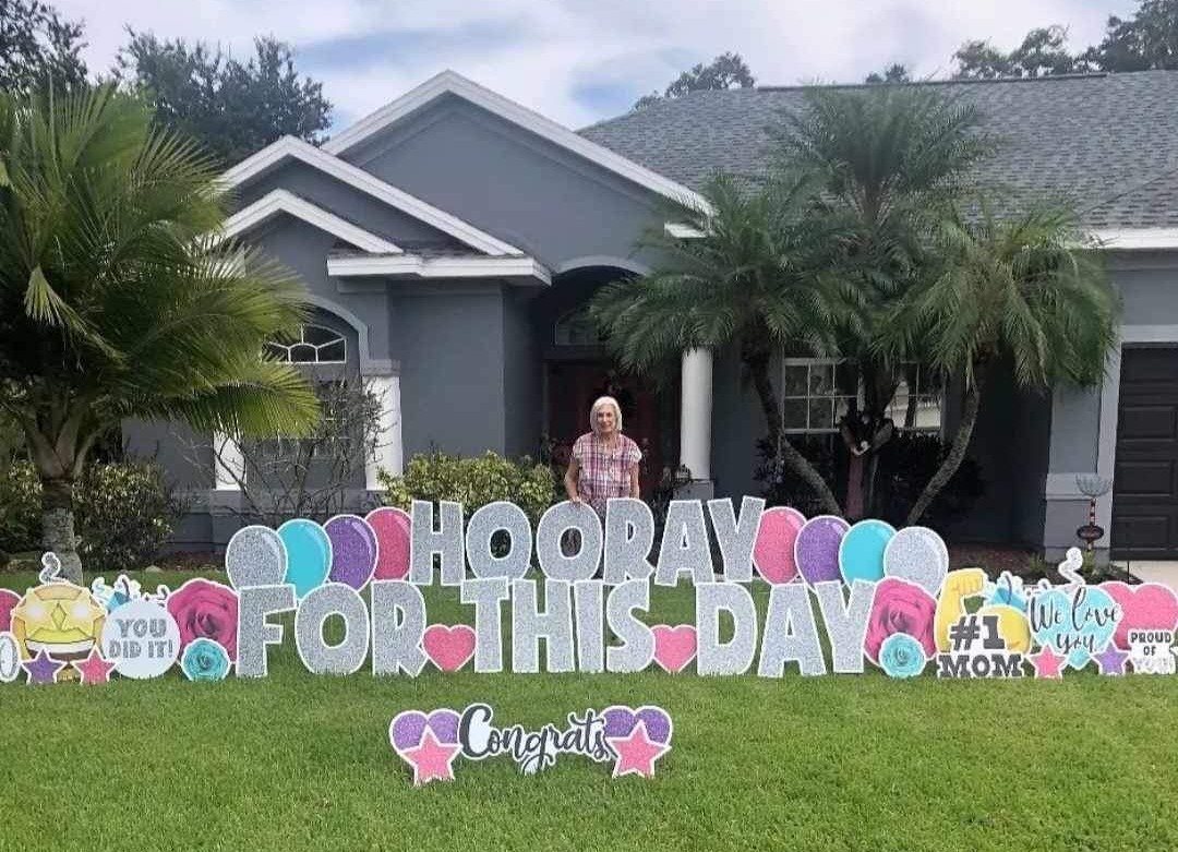 Older woman with "Hooray for this Day Yard sign + other signs - balloons, We Love You, #1 Mom, Proud of You, You Did It, Congrats - sparkling/glitter on all signs - in grays, purple, turqouise, pink, and yellow.  Yard Love Brevard - Lindsey Yoder Owner