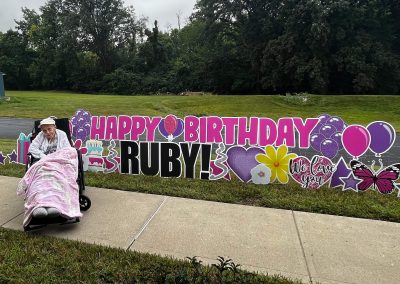 Happy Birthday Ruby - Assisted Living Birthday - Bright Pink, Purple and black, hearts, balloons, butterflies, cake, presents, stars and flowers - with Ruby in her wheelchair in a white cap with a lap blanket