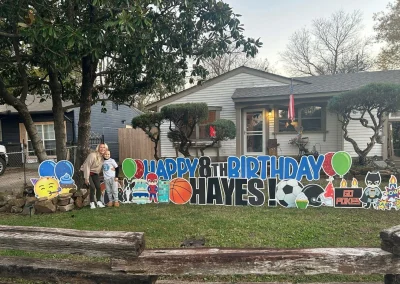 A mother and young son posing to the left of a YL Broken Arrow sign - "Happy 8th Birthday, Hayes!" Superhero and sports symbolism, "Go Pokes", balloons and cupcakes